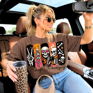 Puede incluir: Una camiseta marrón de manga corta con un estampado gráfico de tres esqueletos sobre fondos de pinceladas verticales naranjas y rosas. El diseño incluye un signo de paz, un esqueleto bailando y un esqueleto bebiendo café con el texto Stay Creepy.