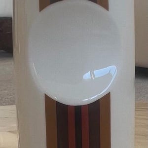 Peut inclure: Cendrier en céramique blanche avec une indentation incurvée et des rayures verticales dans les tons marron, rouge et noir. Le cendrier a une forme cylindrique et une finition brillante, adapté à la décoration intérieure.