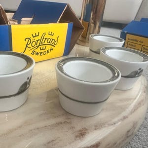May include: Four white ceramic egg cups with a gray rim and a gray floral design. The egg cups are on a marble-like surface. A box with the text "Rörstrand SWEDEN" is in the background.