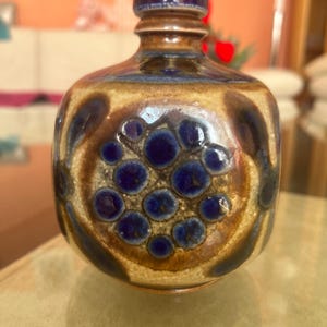 May include: A ceramic bottle with a dark blue stopper and a rounded body. The bottle features a brown and tan base with dark blue circular patterns. The design is reminiscent of mid-century modern pottery.