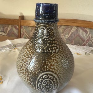 Puede incluir: Jarra de gres con tapa azul oscuro y diseño de rostro. La jarra tiene un cuerpo moteado marrón y beige con un emblema decorativo cerca de la base. La cara tiene barba y está centrada en la jarra.