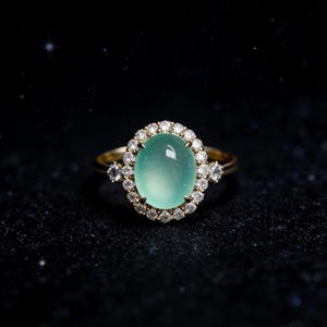 Puede incluir: Un anillo de oro con una gran piedra preciosa ovalada de color verde claro, rodeada por un halo de pequeños diamantes transparentes. Se engarzan diamantes adicionales a ambos lados de la piedra principal. El anillo se muestra sobre un fondo oscuro.