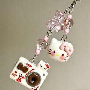 Puede incluir: Un charm blanco y rosa con una cámara y una cabeza de Hello Kitty. La cámara tiene una lente marrón y una pequeña figura de Hello Kitty en la parte superior. Cuentas rosas en forma de corazón y lazo están ensartadas en una cadena plateada.