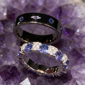 Puede incluir: Dos anillos exhibidos sobre una superficie púrpura. El anillo superior es negro con piedras preciosas azules. El anillo inferior es plateado con piedras preciosas azules y diamantes blancos. Los anillos son probablemente para compromiso o moda.
