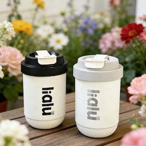 May include: Two white travel mugs with black and gray lids. The mugs have the word "ialu" printed vertically in black on the side. The background features a variety of colorful flowers.