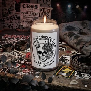 Puede incluir: Una vela encendida en un tarro de vidrio con la etiqueta "Hello darkness my old friend". La etiqueta presenta un diseño de calavera, mariposa y flores. La vela está sobre una superficie de madera con accesorios de temática punk rock.