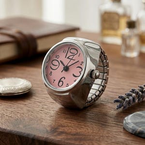 May include: A silver-toned ring watch with a pink face and black numerals. The watch has a stretch band and is set on a wooden surface. A closed silver locket and a brown leather-bound book are in the background.