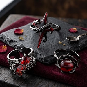 May include: Three gothic-style rings with red and black accents. One ring features a red gemstone, another has a starburst design, and the third has a large red stone. The rings are displayed on a dark surface.