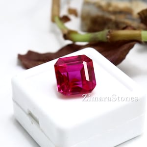 May include: A vibrant, square-cut, deep pink gemstone with a faceted surface that reflects light. The gemstone is positioned on top of a small, white, square plastic display box featuring the text ZimaraStones printed in a simple font.