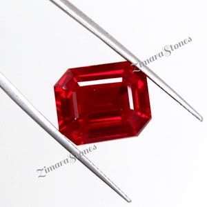 Könnte beinhalten: Ein facettierter, rechteckig geschliffener Rubin-Edelstein, der von einer silbernen Pinzette gehalten wird. Der Edelstein hat eine tiefe, leuchtende rote Farbe mit sichtbaren inneren Reflexionen. Der Text "Zimara Stones" ist sichtbar.