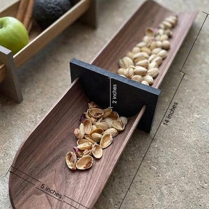 Puede incluir: Una bandeja de madera oscura de 35,5 cm de largo y 12,7 cm de ancho, llena de pistachos. Un divisor negro mide 5 cm de alto. Otra bandeja con frutas y especias está en el fondo.