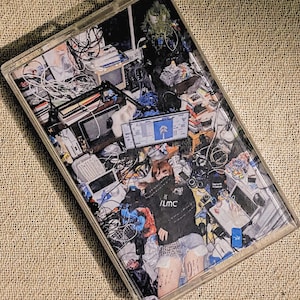 May include: A clear plastic cassette tape case featuring a graphic insert of a person wearing a black hoodie with the text ILMC, surrounded by a cluttered room filled with computer monitors, tangled electronic cables, and various electronic devices.