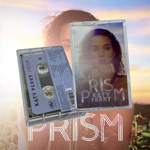 Puede incluir: Una cinta de casete transparente con la portada del álbum "Prism" de Katy Perry. La cinta de casete es visible, con el título del álbum y el nombre del artista impresos en la etiqueta. El fondo presenta una imagen borrosa de Katy Perry.
