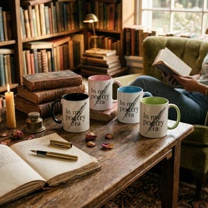 Puede incluir: Cuatro tazas de cerámica con el texto "In my poetry era" y una ilustración de pluma. Las tazas tienen detalles en negro, rosa, azul y verde. Las tazas están sobre una mesa de madera con un libro abierto, un bolígrafo y pétalos de rosa secos.