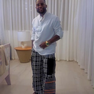 May include: A white long-sleeved shirt with a mandarin collar and loose-fitting trousers featuring a black and white grid pattern, black side panels, and horizontal orange and grey striped accents at the bottom hem, paired with black sandals.