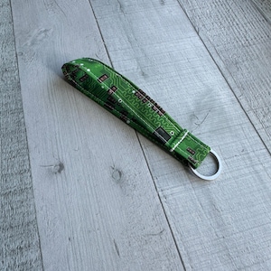 May include: A green fabric keychain wristlet with a circuit board pattern. The wristlet has a silver metal key ring attached. The fabric is a bright green with a white stitched border.