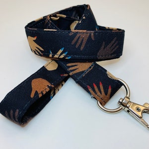 May include: Black lanyard with a repeating pattern of brown hands with colorful nail polish. The lanyard has a silver swivel clip.
