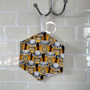 May include: A hexagonal kitchen pot holder with a black background and a pattern of yellow honey jars with white lids. The pot holder is hanging from a hook on a white tiled wall.