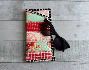Könnte beinhalten: Ein buntes, gestepptes Brillenetui mit Patchwork-Design in roten, rosa- und grünen Blumenmustern. Eine schwarze Sonnenbrille liegt darauf. Das Etui hat eine schwarz-weiße Bordüre.