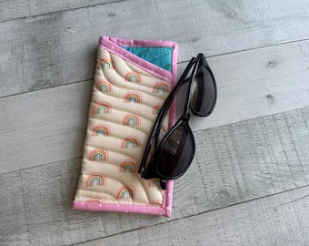 Könnte beinhalten: Ein gestepptes Brillenetui mit Regenbogenmuster und eine schwarze Sonnenbrille. Das Etui ist beige mit bunten Regenbogenmotiven und rosa Einfassung. Die Sonnenbrille ist schwarz und hat ein klassisches Design.