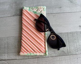 Könnte beinhalten: Eine schwarze Sonnenbrille mit rundem Rahmen liegt auf einem gestreiften, gesteppten Brillenetui. Das Etui hat ein Muster aus weißen und korallenfarbenen Streifen mit einem grünen Blumenrand. Die Sonnenbrille hat dunkle Gläser.