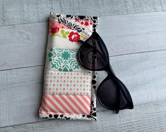 Könnte beinhalten: Eine schwarze Sonnenbrille liegt auf einem farbenfrohen, gesteppten Brillenetui. Das Etui hat ein Patchwork-Design mit floralen, geometrischen und gestreiften Mustern in Rosa, Grün und Weiß. Das Etui ist rechteckig und hat einen schwarz-weißen Blumenrand.