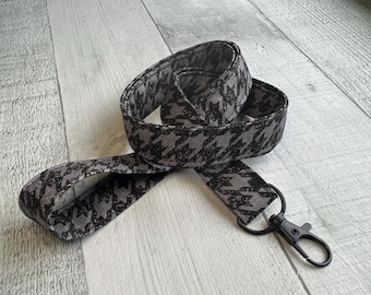 Op de afbeelding: Een stoffen keycord met een herhalend zwart en grijs pied-de-poulepatroon. De band is gemaakt van geweven textiel en is aan één uiteinde voorzien van een matzwarte metalen karabijnhaak voor het bevestigen van diverse voorwerpen.