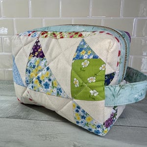 Puede incluir: Una bolsa de cremallera acolchada de tela con estampado floral y asa azul claro. La bolsa está hecha de tela de patchwork con una variedad de patrones florales.