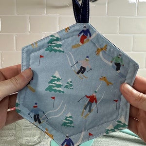 May include: A light blue, hexagonal pot holder with a navy blue trim and hanging loop. The fabric features a repeating pattern of skiers, trees, and red flags on a snowy mountain. The pot holder is held up by a navy blue loop.