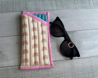 Könnte beinhalten: Ein gestepptes Brillenetui mit Regenbogenmuster und eine schwarze Sonnenbrille. Das Etui ist cremefarben mit Reihen von Regenbogenbögen und einem rosa gepunkteten Rand. Die Sonnenbrille hat einen schwarzen Rahmen und ein goldfarbenes Ringdetail.