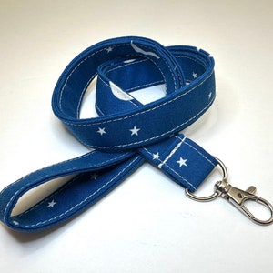 May include: A navy blue lanyard with a white star pattern and white stitching. The lanyard has a silver-colored metal clasp and a loop for attaching keys or an ID badge. The fabric is a sturdy cotton material.
