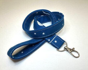 May include: A navy blue lanyard with a white star pattern and white stitching. The lanyard has a silver-colored metal clasp and a loop for attaching keys or an ID badge. The fabric is a sturdy cotton material.