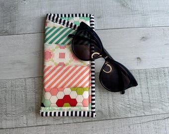 Könnte beinhalten: Gestepptes Brillenetui mit Patchwork-Design in Rosa, Grün und Weiß, akzentuiert mit schwarz-weiß gestreiften Zierleisten. Schwarze Sonnenbrille liegt oben auf dem Etui.