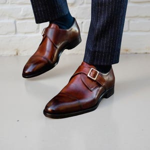 May include: A pair of polished brown leather monk strap shoes featuring a single silver-toned metal buckle closure across the top. The shoes have a smooth, burnished finish with a rounded toe and are worn with dark pinstriped trousers and socks.