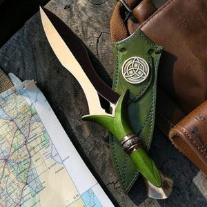 Puede incluir: Una daga artesanal con una hoja de dos tonos y un mango verde. La daga está en una funda de cuero verde con un emblema de nudo celta. Un mapa y accesorios de cuero sugieren aventura y artesanía.