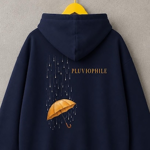 Può includere: Una felpa con cappuccio blu navy con una grafica sul retro che raffigura un ombrello giallo sotto la pioggia. Il testo PLUVIOPHILE è stampato in lettere dorate sopra l'ombrello. La felpa è esposta su una gruccia giallo brillante.