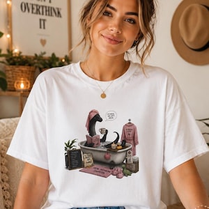 Può includere: T-shirt bianco con un'illustrazione di una creatura nera in una vasca da bagno, che legge e beve. Sullo sfondo, una stampa incorniciata con la scritta "Don't Overthink It". La maglietta è abbinata a jeans blu.