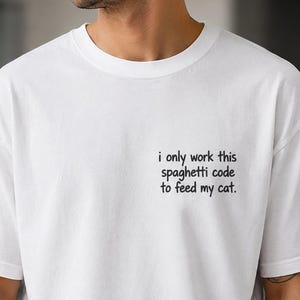 Può includere: Una t-shirt bianca a maniche corte con testo nero sulla parte superiore sinistra del petto che recita: "i only work this spaghetti code to feed my cat." La maglia presenta un girocollo e una vestibilità standard in tessuto bianco.