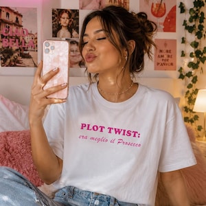 Puede incluir: Camiseta blanca con el texto rosa "PLOT TWIST: era meglio il Prosecco". La persona lleva vaqueros azul claro y sostiene un teléfono rosa. Un letrero de neón que dice "good vibes" está en el fondo.