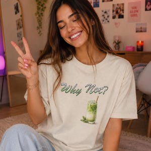 Può includere: T-shirt beige con la scritta verde "Why Not?" e un'illustrazione di una bevanda al matcha. La persona che indossa la maglietta sorride e fa il segno della pace.