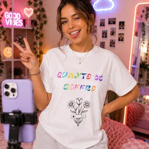 Può includere: T-shirt bianco con la scritta italiana "Quanto ci soffro" in colori arcobaleno sopra una grafica floreale con una benda. La persona indossa pantaloni della tuta grigi e fa il segno della pace.