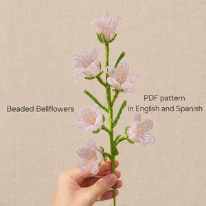May include: A stem of beaded bellflowers with light pink petals and green stems. The text on the image reads: Beaded Bellflowers, PDF pattern in English and Spanish. The flowers are constructed from small beads arranged in a bell-like shape.