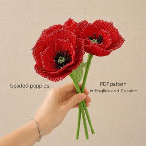 Op de afbeelding: Twee handgemaakte kralenklaprozen met levendige rode bloemblaadjes en donkere centra, voorzien van groene stelen. De tekst op de afbeelding luidt: beaded poppies, PDF pattern in English and Spanish. Deze decoratieve bloemen zijn gemaakt van kleine kralen.