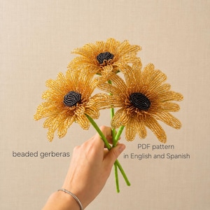 May include: Three handcrafted beaded gerbera flowers featuring golden yellow petals and dark black centres. The stems are green and slender. The text on the image reads beaded gerberas and PDF pattern in English and Spanish.
