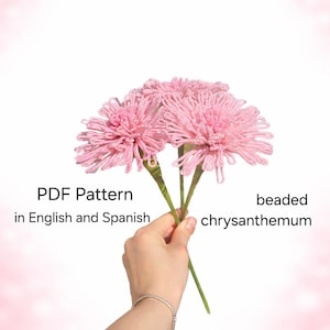 Puede incluir: Tres flores de crisantemo con cuentas rosas, sostenidas por una mano, sobre un fondo rosa suave. La imagen incluye el texto "PDF Pattern in English and Spanish".