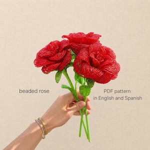 Può includere: Tre vivaci rose rosse realizzate con piccole perline con steli e foglie di perline verdi. Il testo sull'immagine recita: beaded rose, PDF pattern in English and Spanish. Le rose sono disposte in un grappolo con petali stratificati.