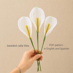 Könnte beinhalten: Drei handgefertigte, perlenbesetzte Calla-Lilien mit weißen Blütenblättern und leuchtend gelben Kolben, die jeweils an einem grünen Stiel befestigt sind. Der Text auf dem Bild lautet beaded calla lilies und PDF pattern in English and Spanish.