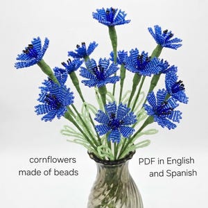 May include: A bouquet of blue beaded cornflowers in a decorative vase. The flowers have green stems and leaves. The vase is a smoky gray color. Text on the image reads "cornflowers made of beads" and "PDF in English and Spanish."