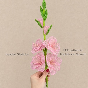 May include: A beaded Gladiolus flower crafted from pink and green glass beads, featuring a tall stem with multiple blossoms and buds. The text on the image reads: beaded Gladiolus, PDF pattern in English and Spanish.