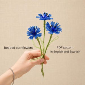 Könnte beinhalten: Drei Kornblumen aus kleinen blauen Perlen mit schwarzen Zentren, befestigt an dünnen grünen Perlenstielen. Der Text lautet: beaded cornflowers PDF pattern in English and Spanish. Die Blumen sind in einem Bündel mit hellgrünen Blättern angeordnet.